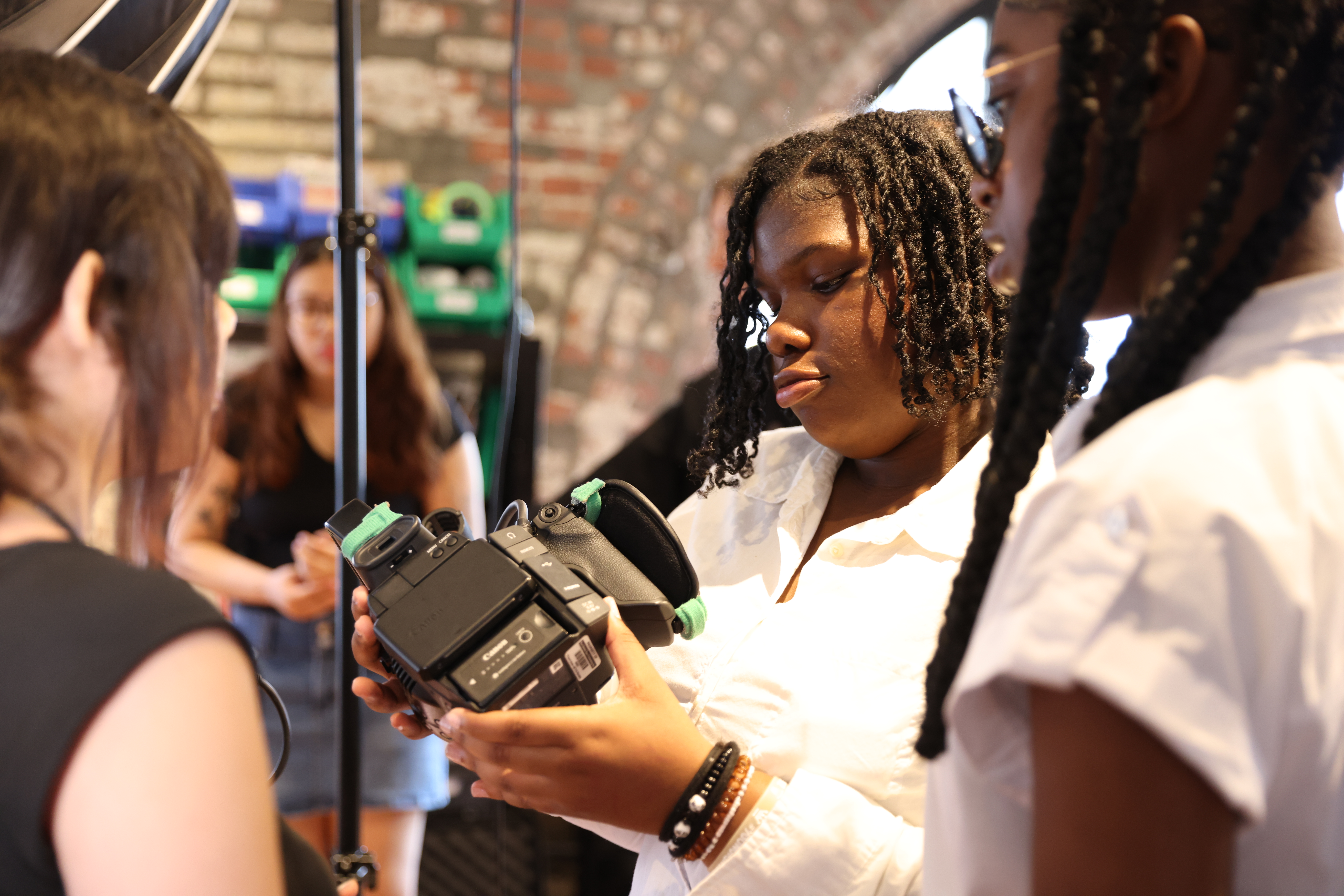 A young student operating a camera. In the foreground, two other students look on. In the background is another student and a shelf of supplies.